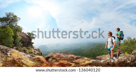 Panorama with two male backpackers standing on top of a mountain and looking to a valley