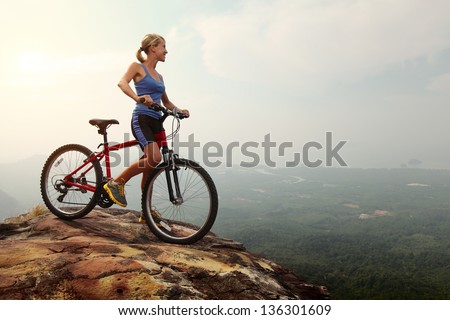 Young slim lady standing on top of a mountain with bicycle and enjoying valley view