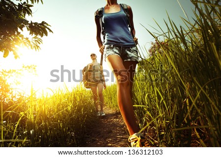 Two young ladies walking with backpacks on a thin path through a lush tropical meadow