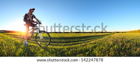 Young Man Cycling On A Rural Road Through Green Spring Meadow During Sunset