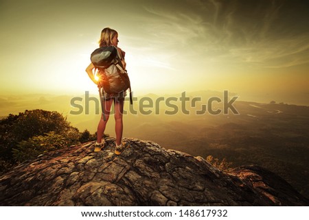 Lady hiker with backpack standing on top of a mountain and enjoying sunrise