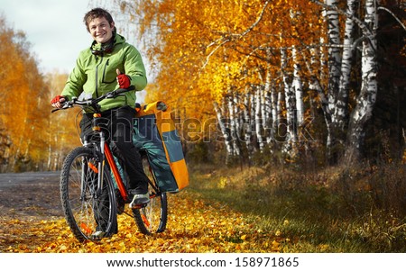 Young happy bicycle tourist with his loaded bike standing on an autumn road in a sunny day