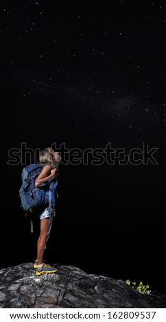 Female hiker with backpack standing on top of a mountain and looking at night sky with stars