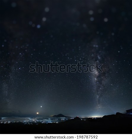Panorama of a night sky with stars and Milky Way on equatorial latitude with mountains and illuminated town below. Tilt shift effect used