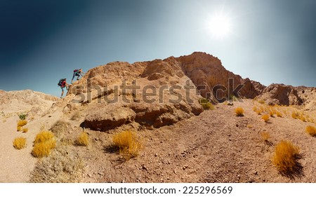 Panorama of two hikers crossing rocky terrain in the desert at sunny day