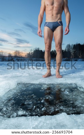 Young man standing by ice hole and ready to swim in the winter water