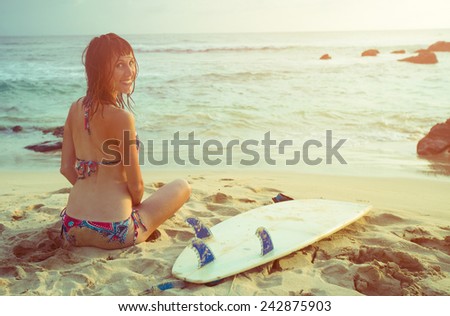 Smiling lady surfer sitting on the sandy beach at sunset
