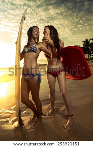Two smiling ladies surfers standing with surf boards on the beach and showing shaka signs