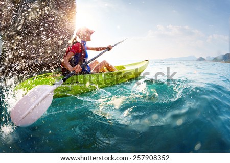 Young lady paddling hard the kayak with lots of splashes near the cliff at sunny day