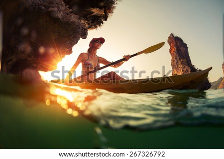 Young lady paddling the kayak in a bay with limestone mountains. Split shot with underwater view at sunset