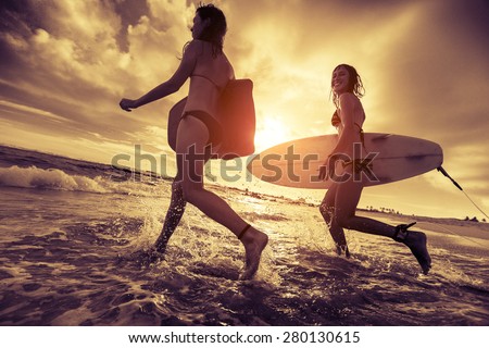 Two ladies surfers running with surfboards into the sea at sunset