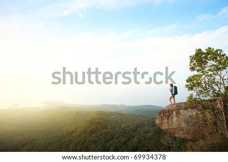Young woman with backpack standing on cliff's edge and looking to a sky Young woman with backpack standing on cliff's edge and looking to a sky