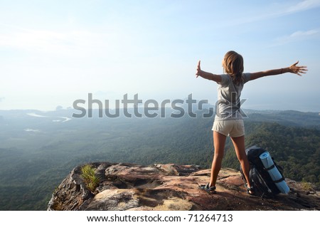 Young woman with backpack standing on cliff\'s edge and looking to a sky with raised hands