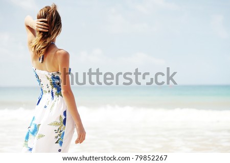 Young woman in summer dress standing by blue sea and looking to horizon