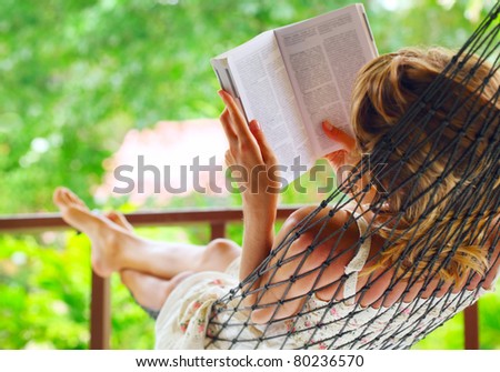 Young woman lying in hammock in a garden and reading a book. Shallow DOF. Focus on a left shoulder
