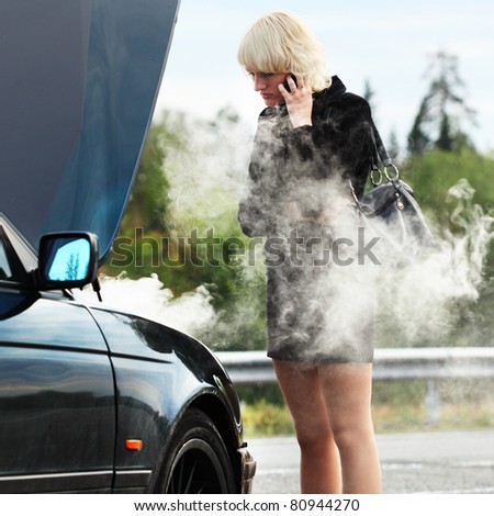 Young blonde woman standing by smoking and broken car and calling with cell phone
