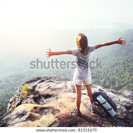 Young woman standing with raised hands with backpack on cliff\'s edge and looking into a wide valley