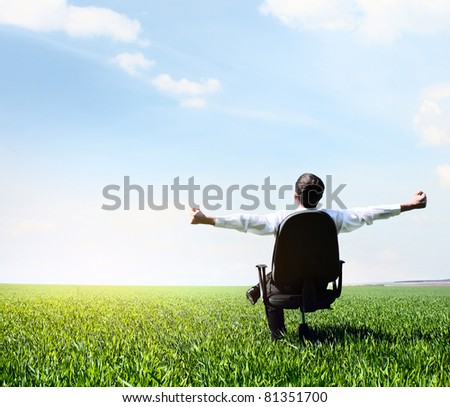 Young man with raised hands sitting in char on green meadow and looking to a sky with clouds