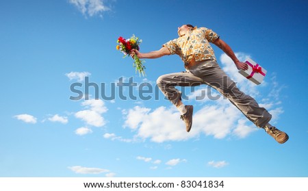 Young happy man jumping with flowers and gift box on blue sky background