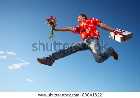 Young smiling man jumping with flowers and gift box on blue sky background