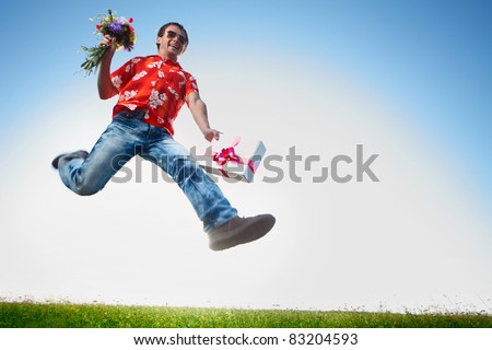 Young happy man in sunglasses jumping with flowers and gift box on blue sky background