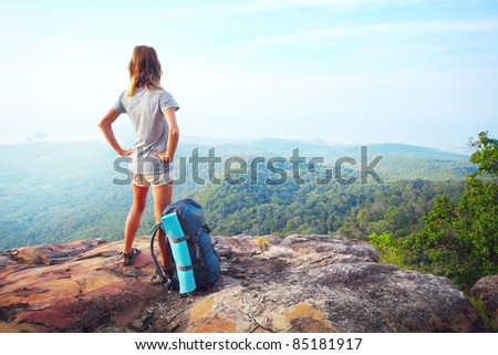 Young woman standing with backpack on cliff\'s edge and looking into a wide valley