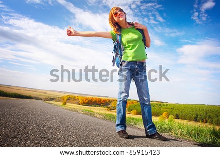 Young smiling woman in glasses and with backpack hitch hiking on an asphalt road