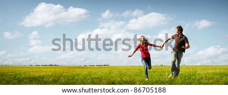 Young happy couple running on a green meadow with blue cloudy sky on the background
