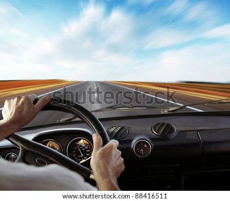 Driver\'s hands on a steering wheel with motion blurred road and sky