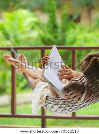Young woman lying in a hammock in garden and reading a book