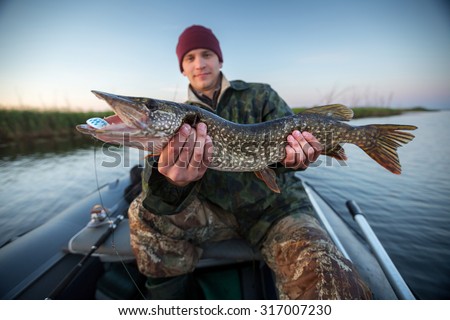 Happy young fisherman holding pike and sitting in the boat on the lake