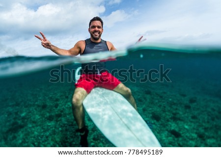 Happy surfer waits the wave on line up with surf board looks at camera and shows the Peace signs