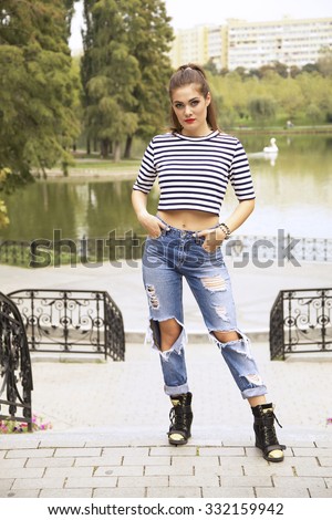 A portrait of a young beautiful glamorous brunette girl in fashionable striped and cropped top and blue jeans in the park in summertime.