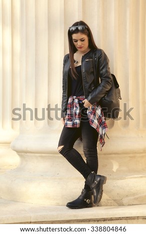 A portrait of a stylish hipster girl in leather black jacket with black leather backpack posing on a building background.