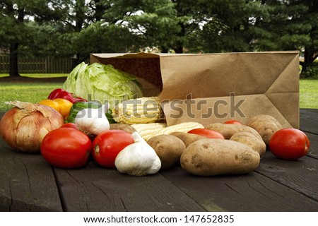 Grocery paper bag on picnic table with assorted vegetables laying around it.