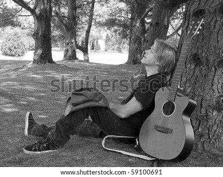 Rocker taking a break outdoors by resting next to large tree with guitar leaning on tree next to him.