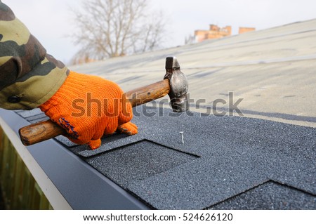Worker hands installing bitumen roof shingles. Worker Hammer in Nails on the Roof. Roofer is hammering a Nail in the Roof Shingles. Construction Nails Vapor barrier and Waterproofing. Unfinished roof.