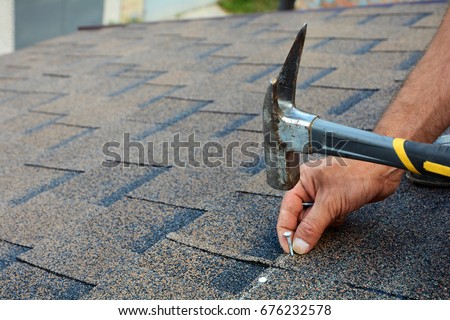 Worker hands installing bitumen roof shingles. Worker Hammer in Nails on the Roof. Roofer is hammering a Nail in the Roof Shingles. Unfinished roof.