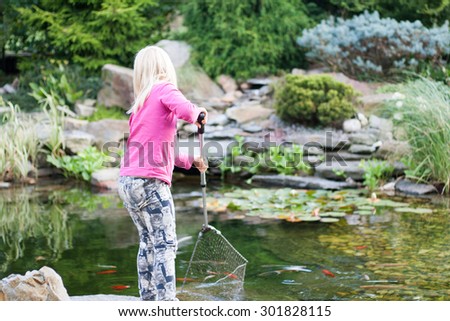 young blonde girl in pink clothes catching fishes into net from pond. Cute little girl