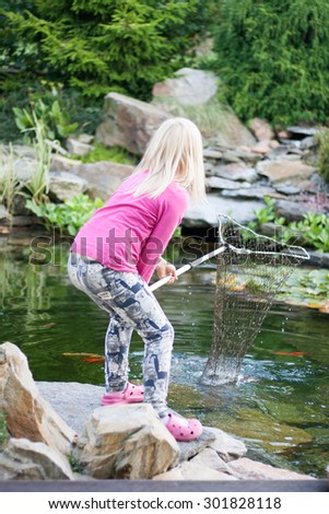 young blonde girl in pink clothes catching fishes into net from pond. Cute little girl