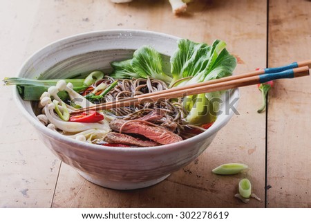 Spicy japanese soup with soba noodles, beef steak, shimeji mushrooms, bok choy, leeks, chili pepper and green onion served in the white oriental bowl on wooden background