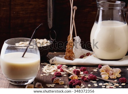 Set of non-dairy milk: oat milk in glass and jug on old rustic wooden background, with oat flakes, sugar sticks, honey and oat biscuits. Dark rustic style.