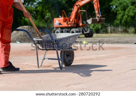Worker with a shovel and a mortar cart. In the background is big excavator. A man is working on a construction site. Photo was taken on a nice sunny day.