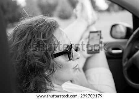 Young attractive girl sitting in her car and holding mobile phone.She is wearing black glasses and white shirt. Mobile phone is black, while coupe cabriolet car is bright blue.Black and white photo.
