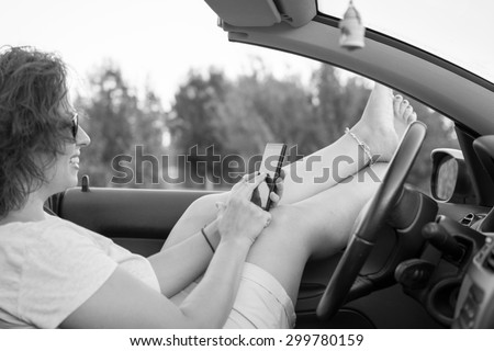 Young attractive girl sitting in her car and holding mobile phone.She is wearing black glasses and white shirt. Mobile phone is black, while coupe cabriolet car is bright blue.Black and white photo.