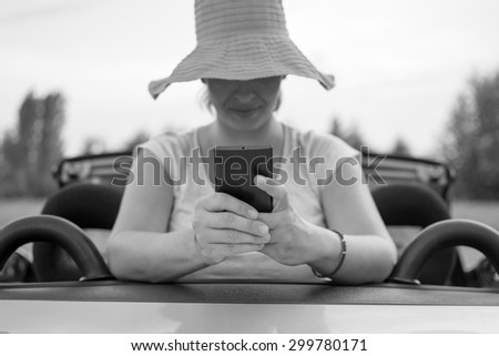 Young attractive girl using mobile phone in her car. She is wearing pink hat and white shirt. Mobile phone is black, while coupe cabriolet car is bright blue. Black and white photo.