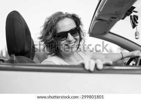 Beautiful young woman in white shirt and black glasses, smiling from a car. Black and white photo.