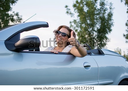 Beautiful young woman smiling and gives thumb up from a car. She is wearing black glasses and white shirt.