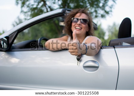 Beautiful young woman in white shirt and black glasses, smiling from a car giving thumbs up.Coupe cabriolet is bright blue.Interior is black-blue color. Focus on the hand and thumb.