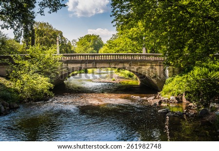 Bridge over peaceful waters - older stone road bridge over a blue stream or brook framed by green summer trees and a patch of puffy blue sky